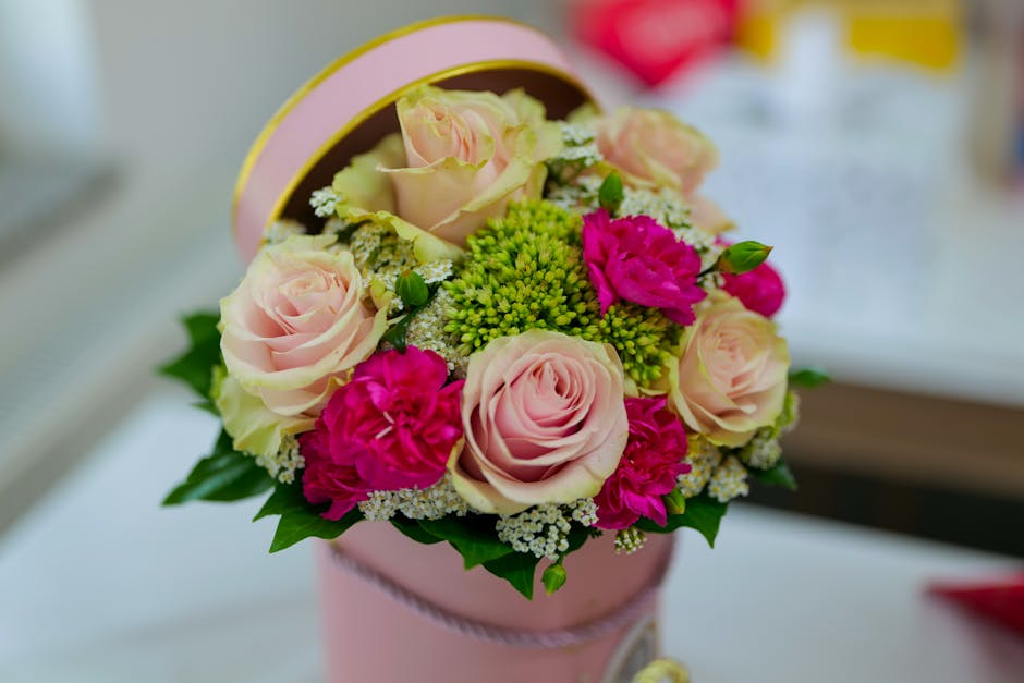 A round, pastel pink floral gift box with a ribbon detail, filled with a bouquet of fresh, pastel pink roses and vibrant magenta carnations, complemented by green foliage and delicate white filler blo