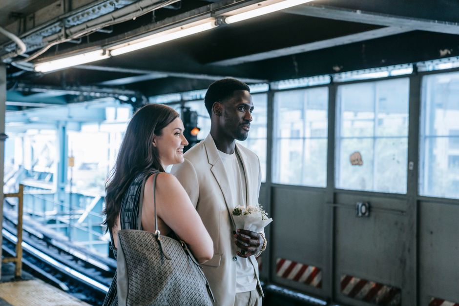 A woman and a man stand on a platform at Ruislip Manor station, illuminated by natural light from large windows behind them. The woman has long dark hair and is smiling, holding a beige handbag over h