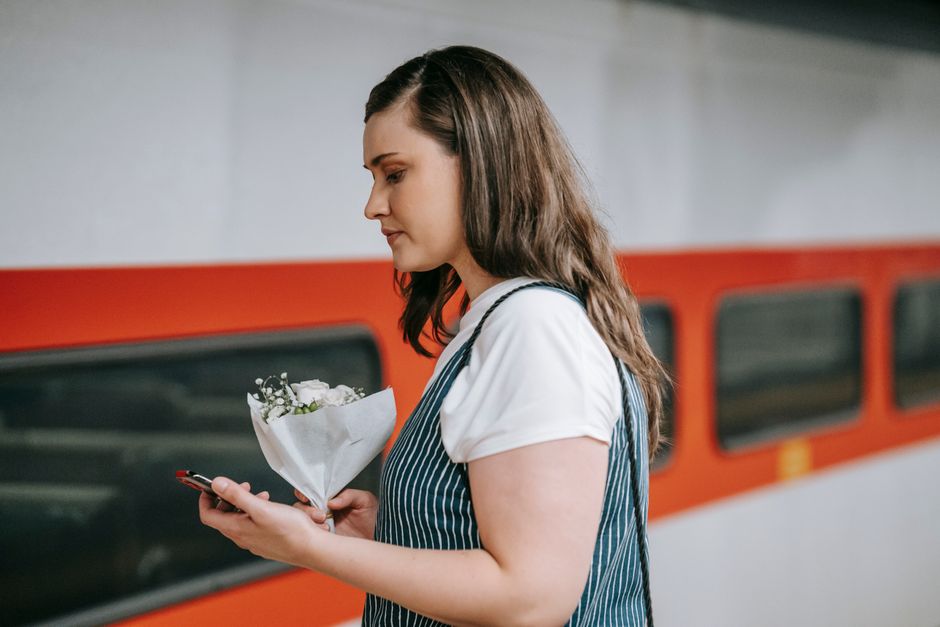 A young woman with long, wavy brown hair stands on a train platform, holding a small bouquet of fresh white flowers wrapped in white paper. She is dressed in a white t-shirt and a blue and white strip