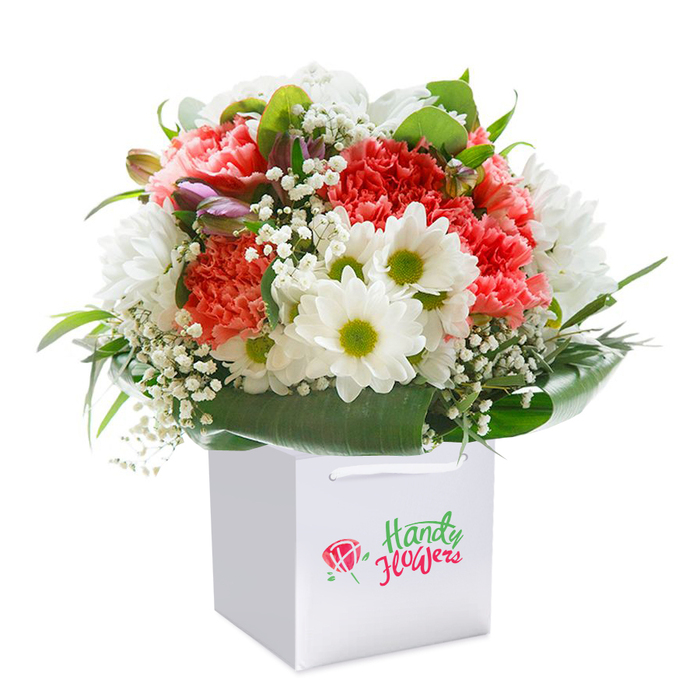 Standing upright within a beautifully structured white gift bag, this lush bouquet shines as a centerpiece of floral artistry-just the sort you'd expect from a boutique florist in South Ruislip. The meticulous arrangement features a vibrant blend of coral and soft peach-toned carnations, their richly ruffled petals overlapping to create an irresistible sense of depth and liveliness. Bright white daisies with distinct yellow-lime centers provide sparkling contrast, while sprays of delicate baby's breath intersperse throughout, imbuing the arrangement with an airy lightness, much like a gentle breeze near the Rayners Lane open spaces. Nestled among the blossoms, subtle purple buds-perhaps alstroemeria or similar-add gentle intrigue, their muted color hinting at promise yet to unfold. Encircling the base, broad, glossy leaves and lush green foliage cradle the bouquet in a vivid wrap, providing substance and visual cohesion. The clean white bag, topped with sturdy ribbon handles, transforms the bouquet into a ready-to-gift token that feels personal and thoughtfully prepared. Whether for birthdays, anniversaries, or sending congratulations, this arrangement radiates the freshness and heartfelt style synonymous with South Ruislip-and stands as a tribute to classic floristry elevated by a sense of place and occasion.